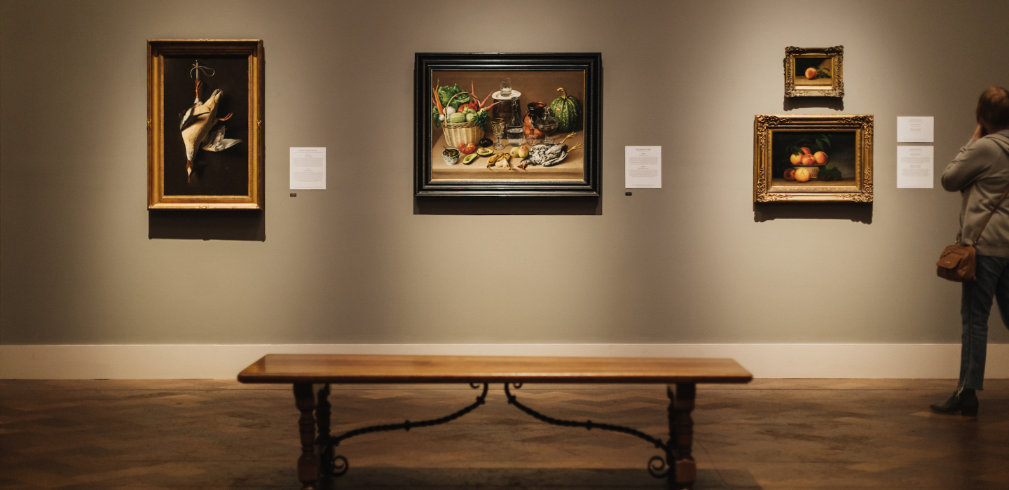 An adult wearing pants and a long sleeve sweater is look at works of still life art on a wall at the San Diego Museum of Art.