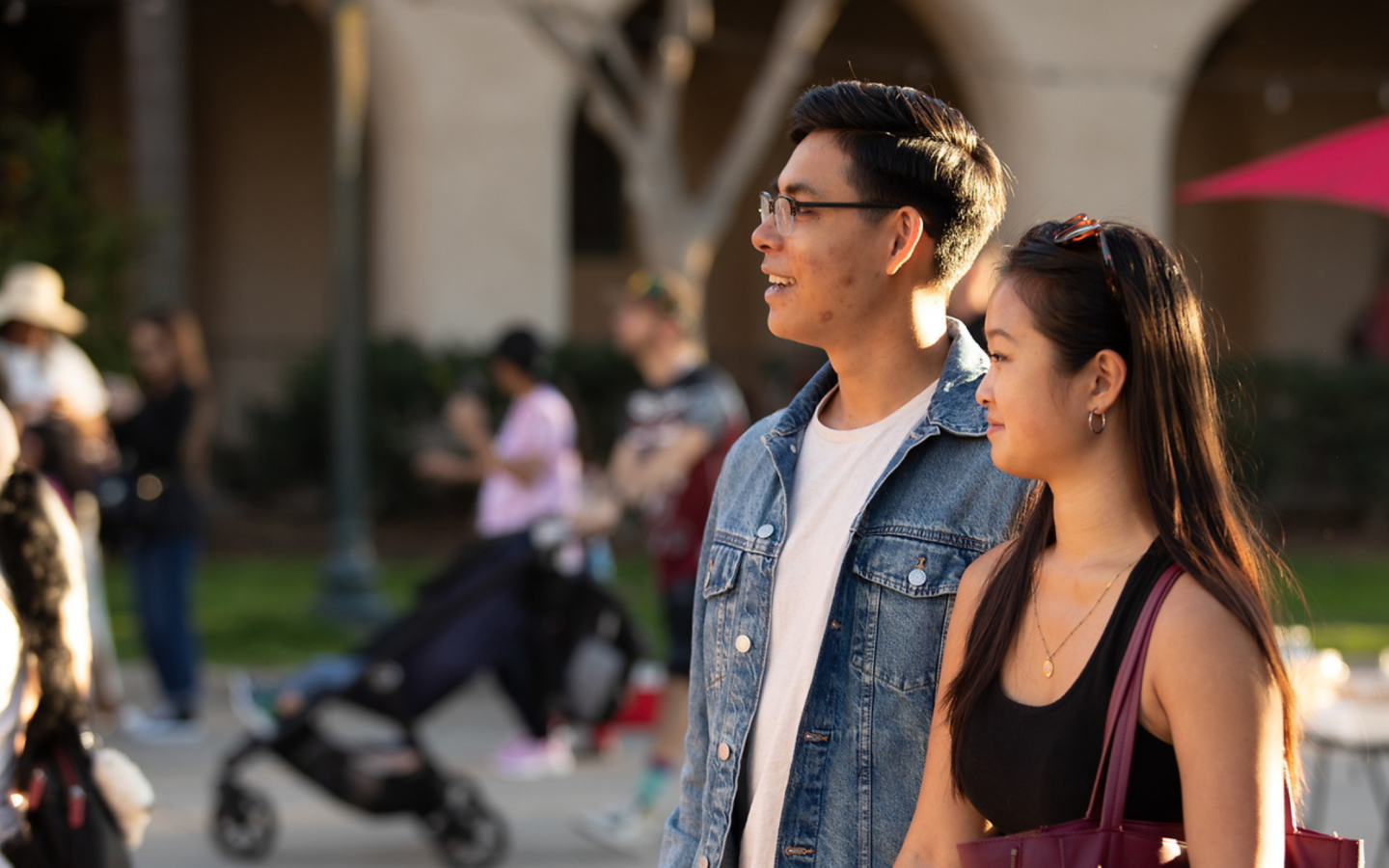 Two young adults pictured on the right looking to the left. There is a person with short brown hair and glasses wearing a white t-shirt and jean jacket. The second person has long brown hair and is wearing a black sleeveless tank top.