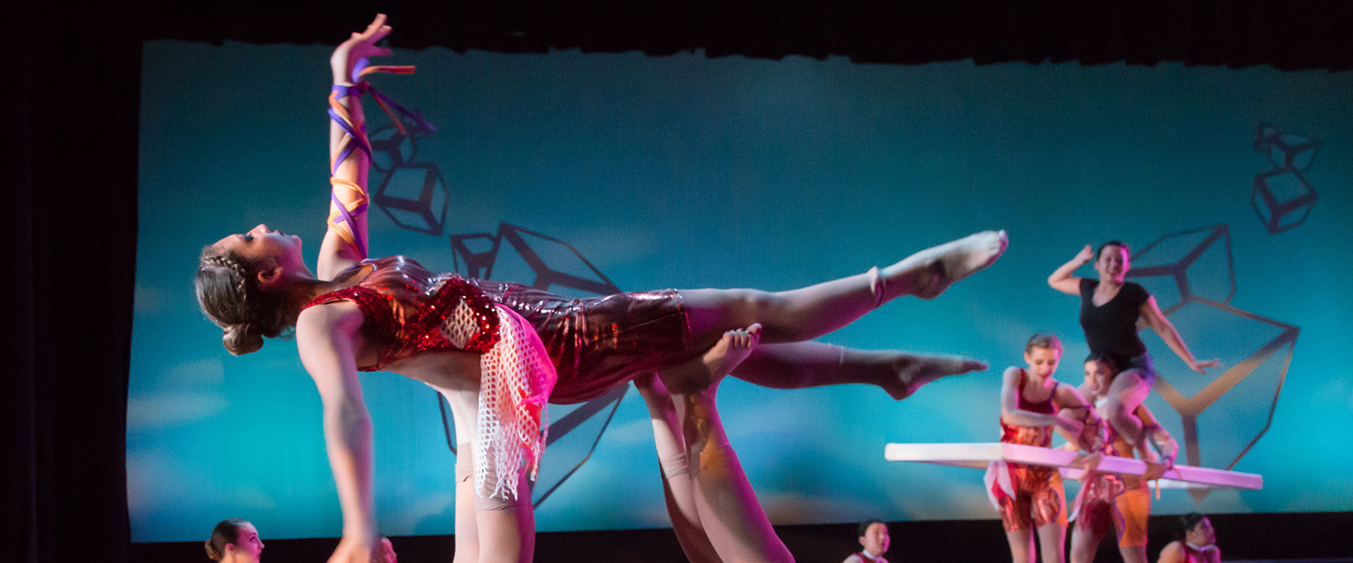 A group of performers wearing brightly colored costumes dancing with one dancer being lifted into the air by two pairs of feet.