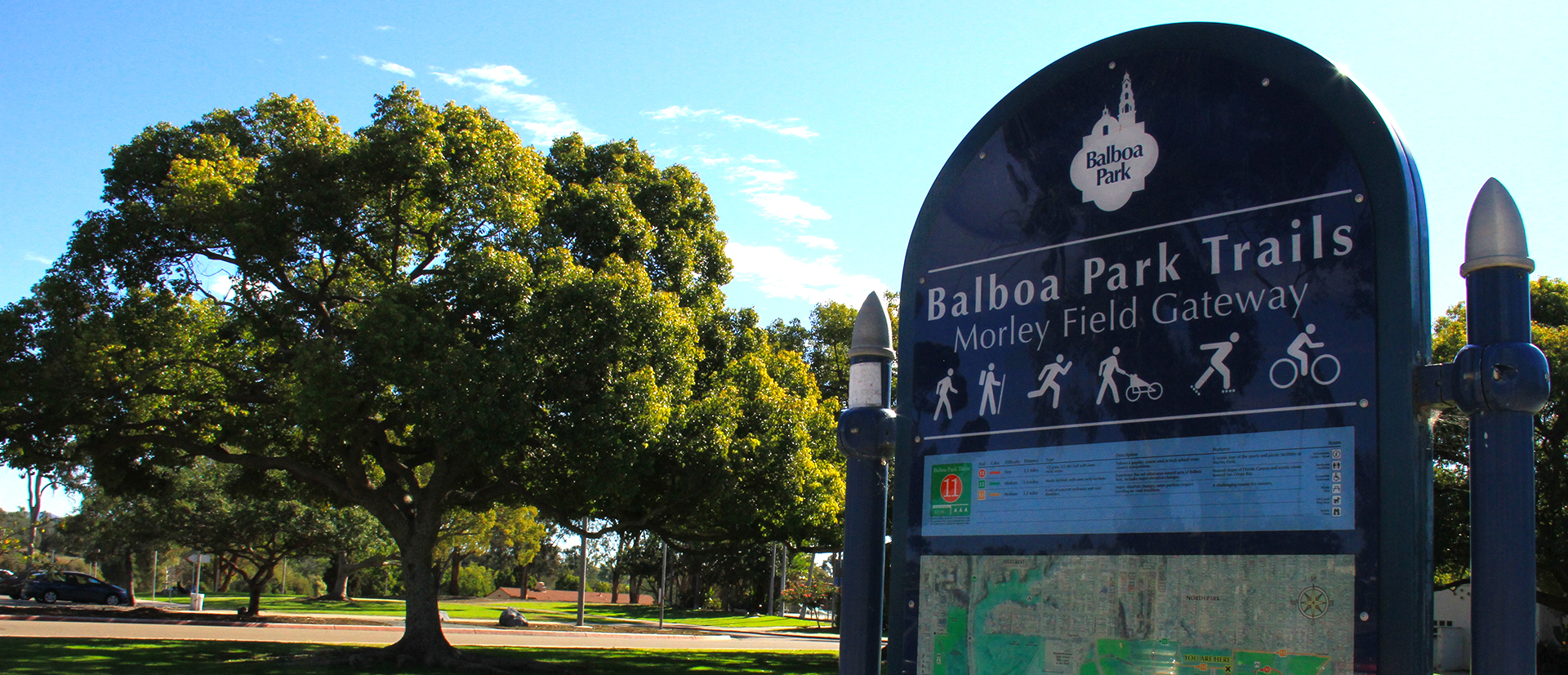 Balboa Park Trails sign for Morley Field Gateway with a trail map and legend. Trees, blue sky and a road are in the background.