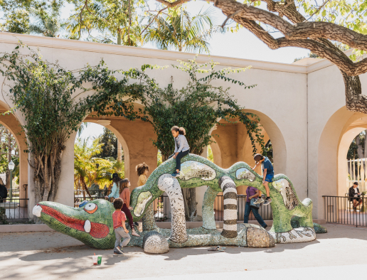 A diverse group of children climbing and playing on the Nikigator sculpture outside the Mingei Museum.