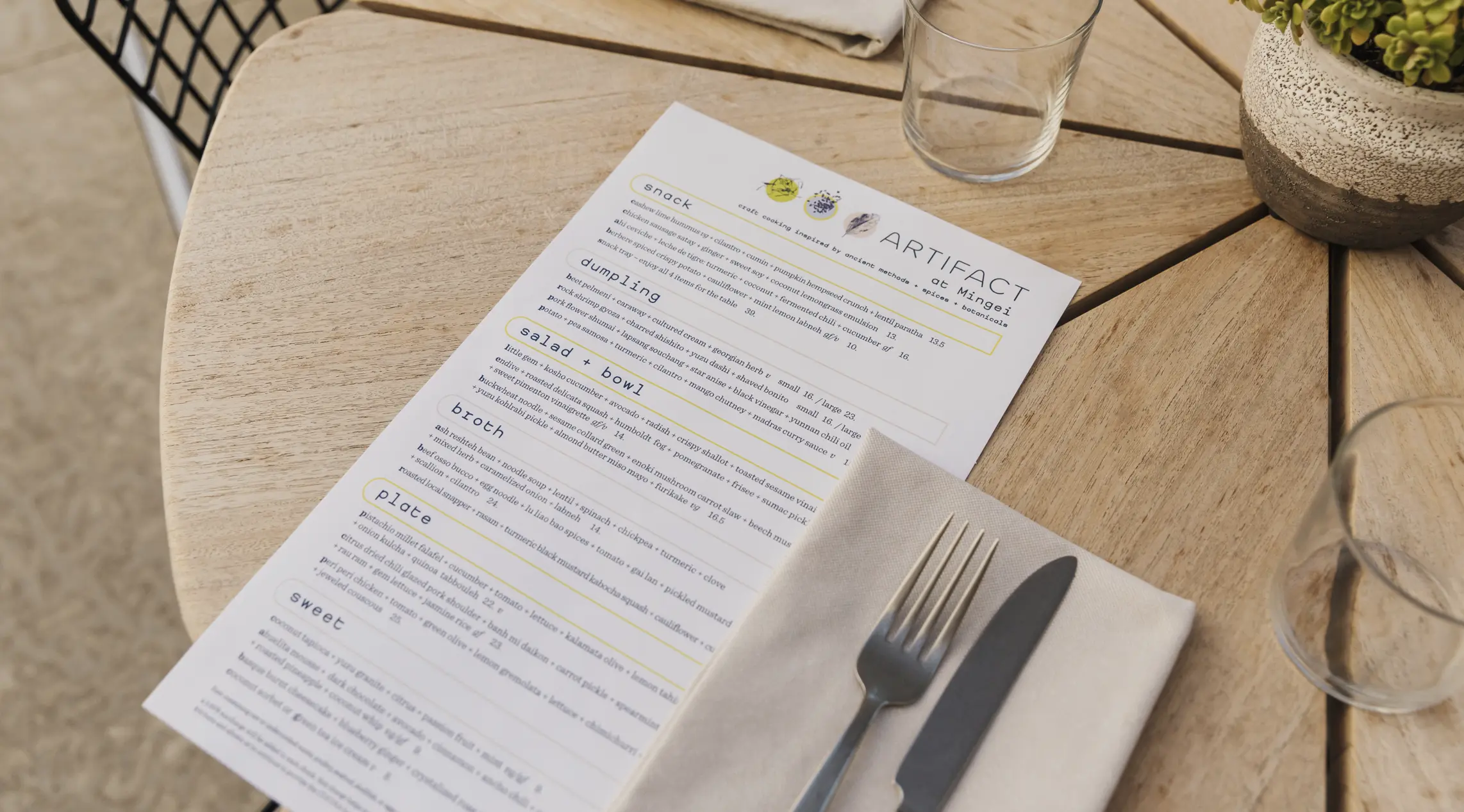 A menu for the restaurant Artifact at Mingei sitting on a round wooden table with utensils, napkins, and drinking glasses surrounding it.