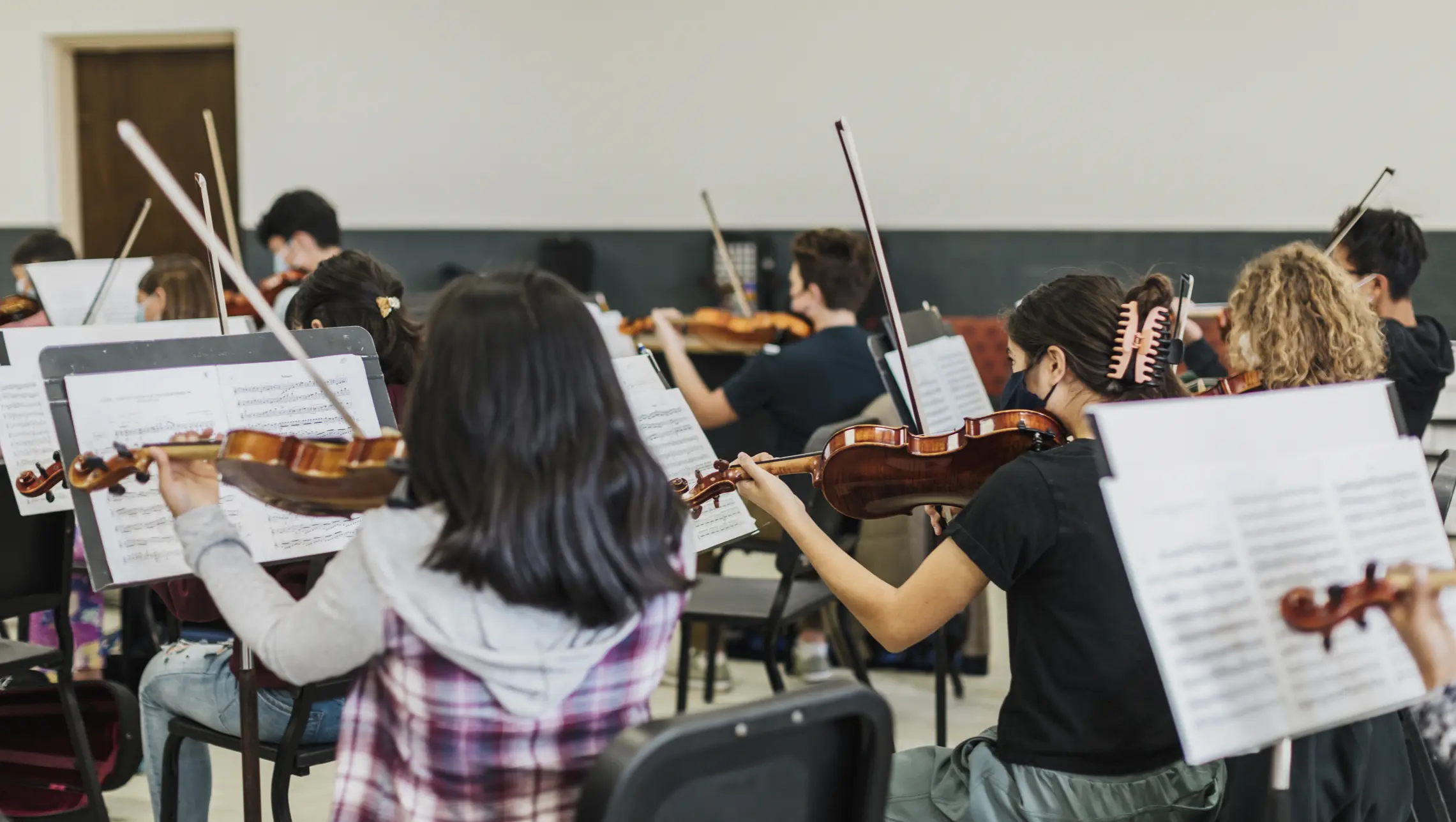 A group of young teenagers playing violins with sheet music on stands in front of them. Many of them can be see wearing face masks.