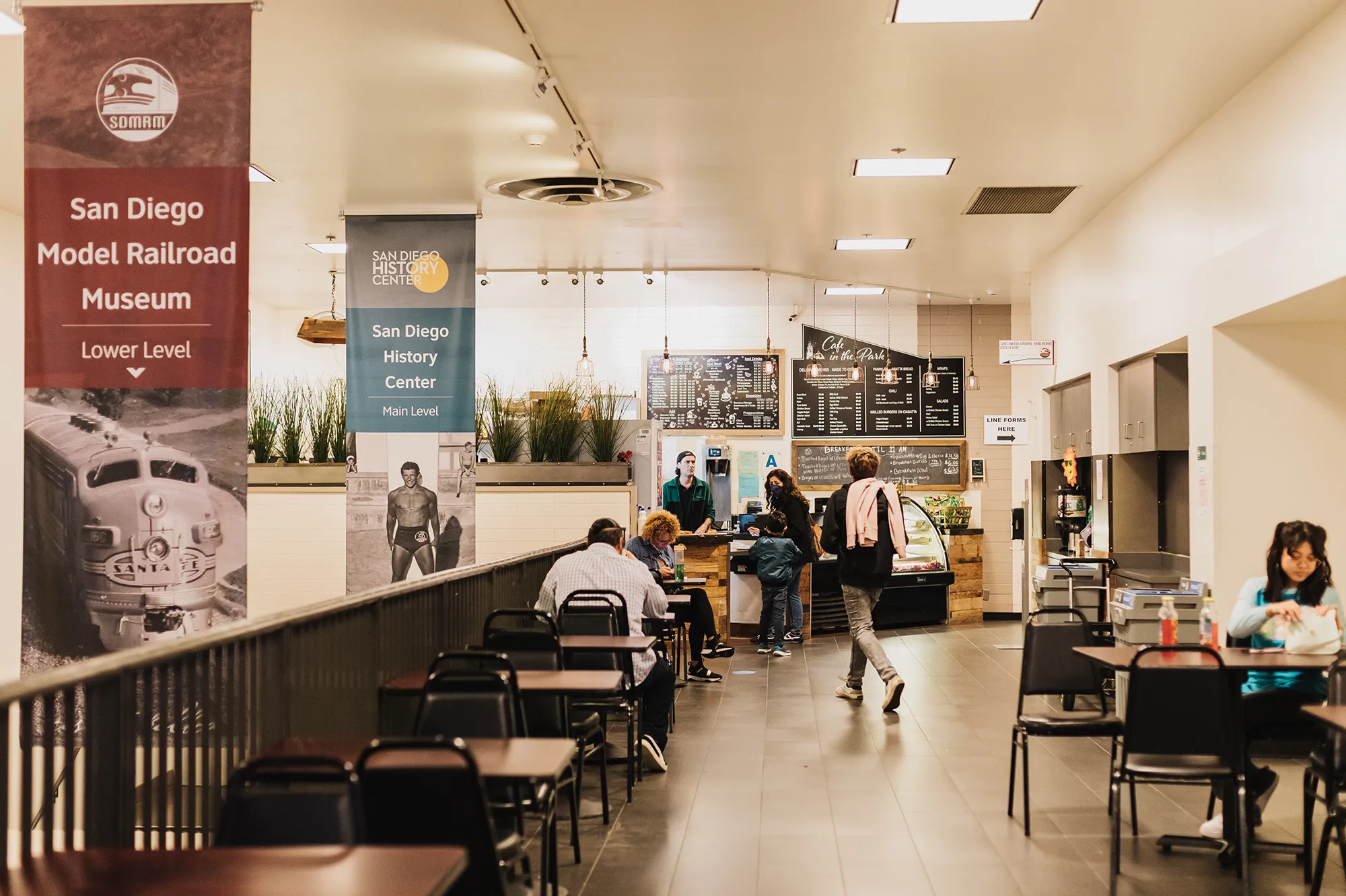 Several people of varying ages and appearance are sitting at Cafe in the Parks dining area and an adult with a child are ordering at the cash register in the background..