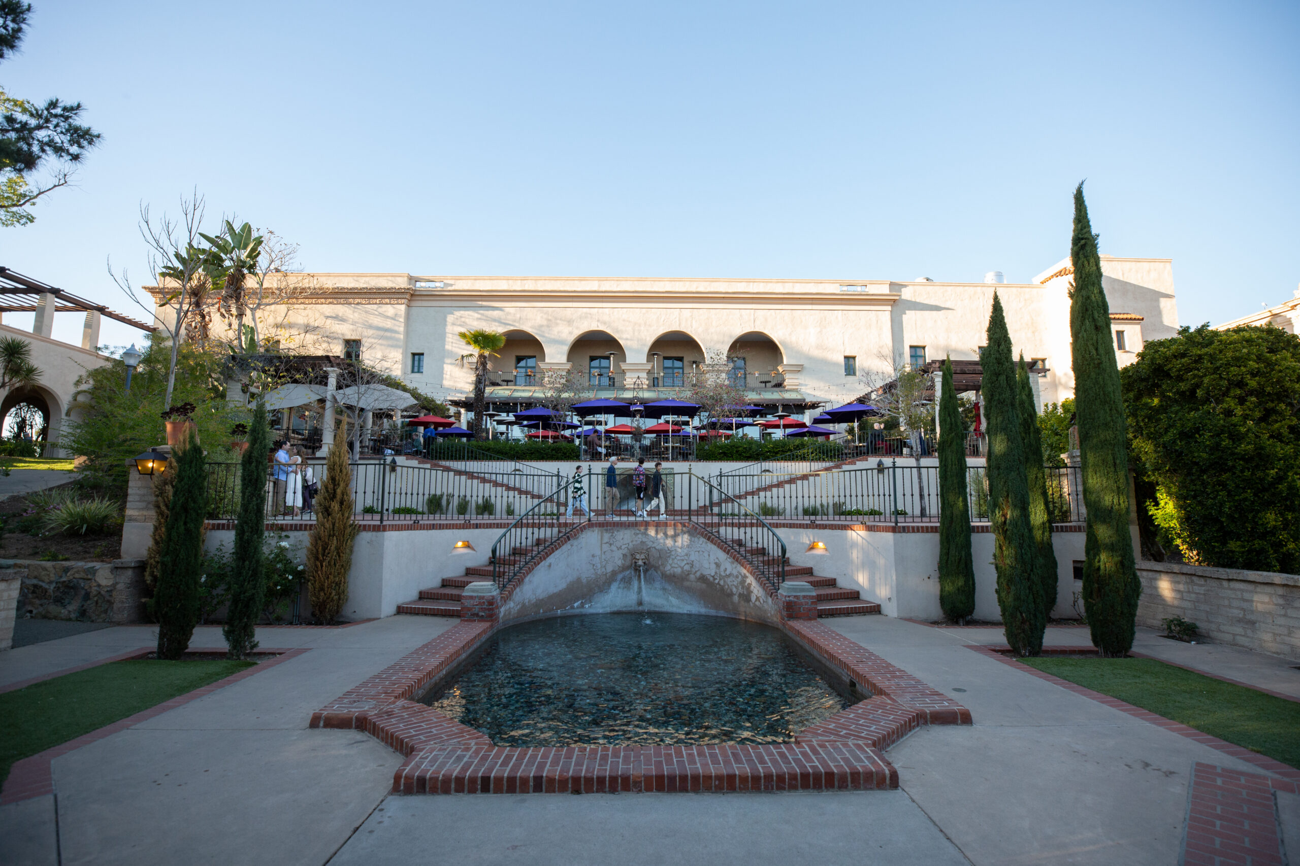 A red brick lined fountain in the center and two sets of red bricked stairs on the right and left hand side leading up to an outdoor patio with tables and red and purple umbrellas.