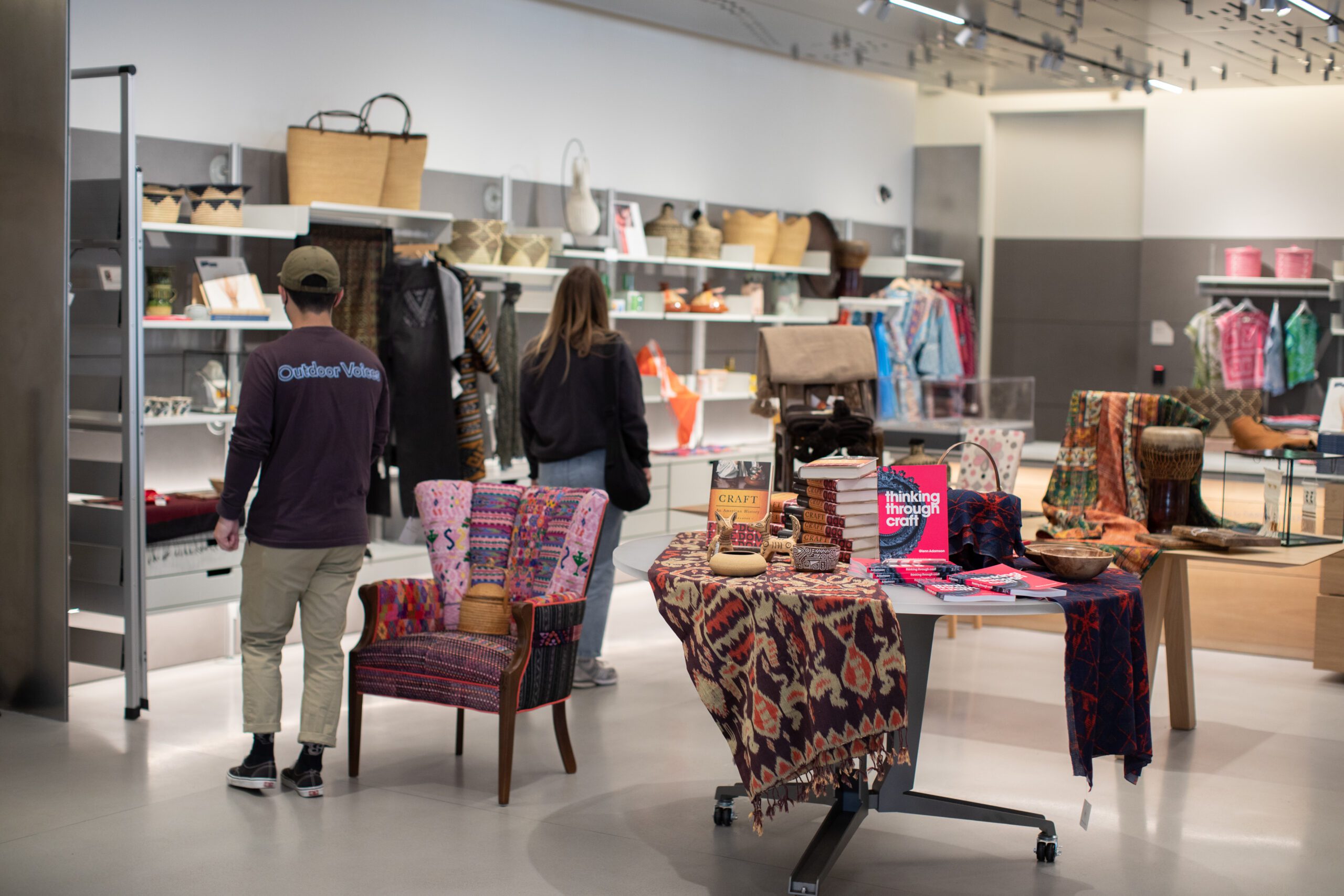 Two people walking through the Mingei gift shop. A variety of clothes, books, bags, and other items are available for purchase.