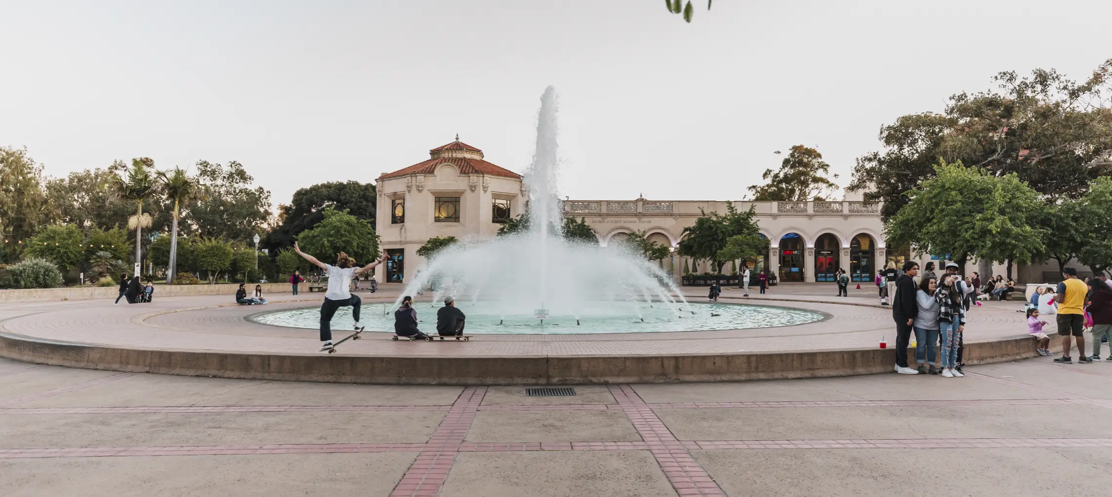 Bea Evenson Fountain in front of the Fleet Science Center. A young group of teenagers are skateboarding on the fountains edge with several other groups of visitors sitting or taking photos nearby.