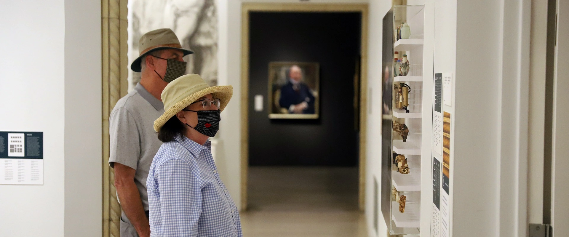 Two adults, one with gray hair and one with short brown hair are wearing masks over their faces and looking at way finding signage in the San Diego Museum of Art.