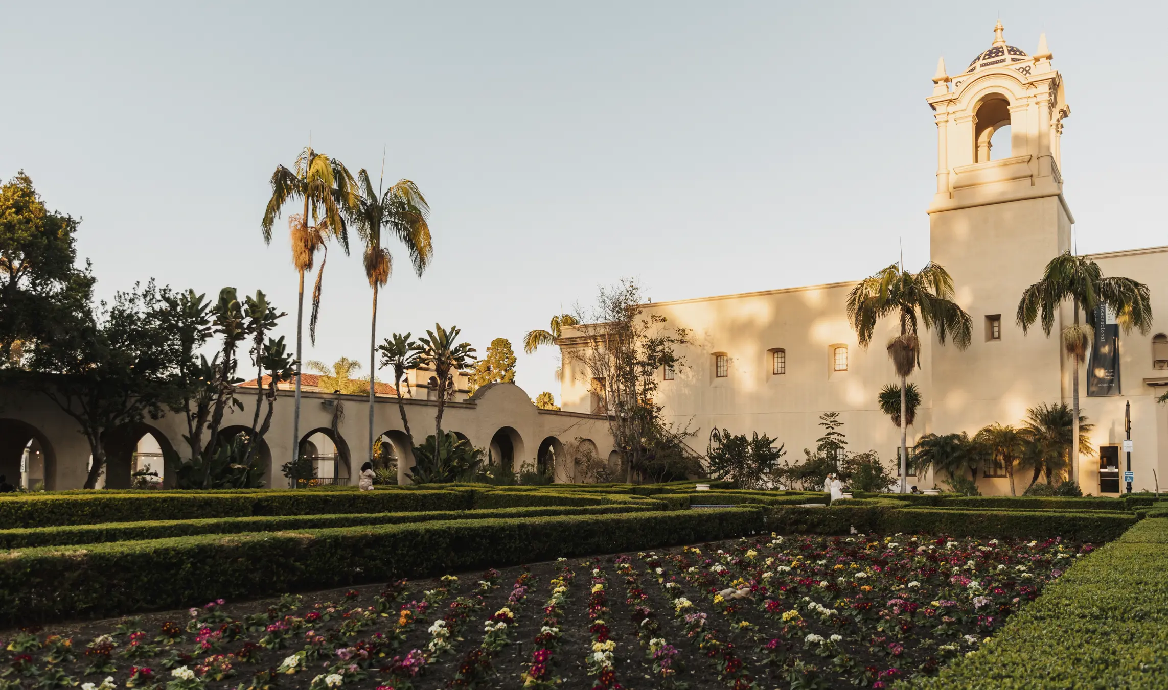 Alcazar garden with rows of flowers and trimmed bushes in the foreground and the back of the Mingei Museum in the background.