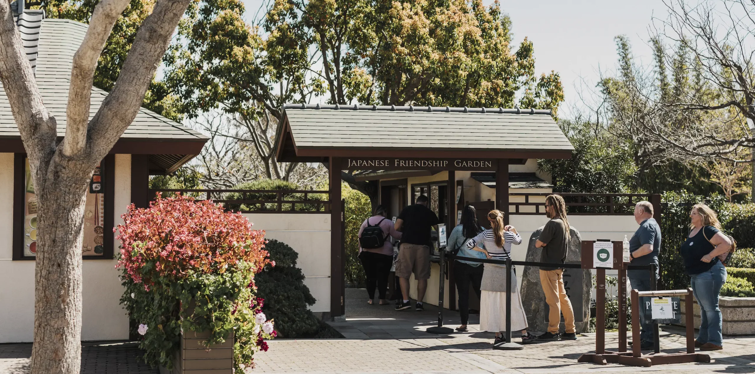 A diverse group of adults waiting in line at the entrance to the Japanese Friendship Garden. A brown and white structure and trees are in the background.