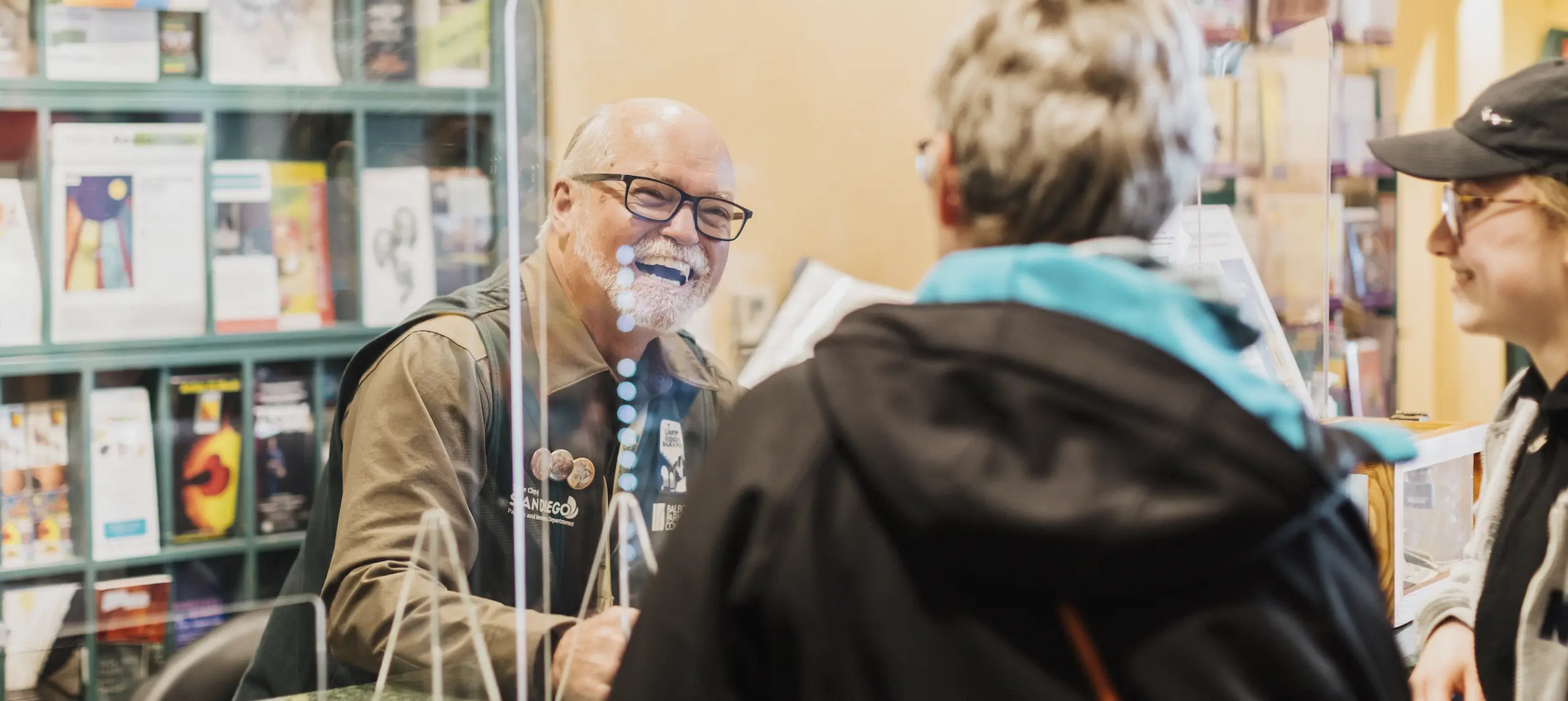 Three adults talking at the Balboa Park Visitors center. Information pamphlets and brochures can be seen in the background.