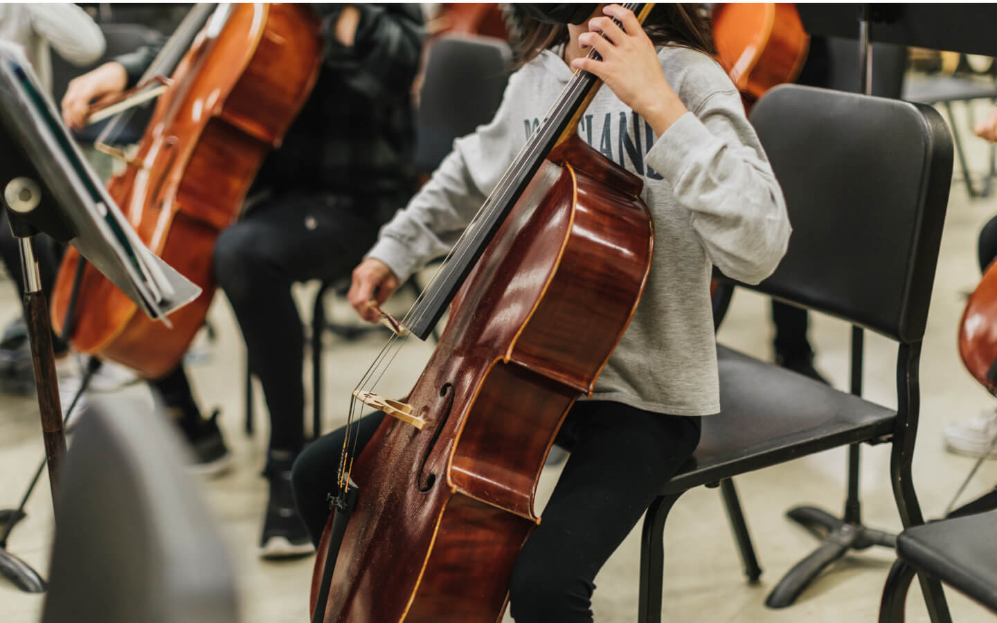 A child with medium brown hair and wearing a gray sweatshirt and black pants is playing a sheet of music with a cello while seated in a black chair.