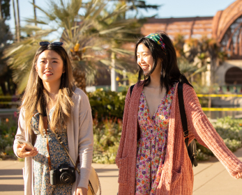 Two young adults with long brown hair. They are walking with the lily pond and botanical building in the background. One adult is wearing a peach colored sweater with a multicolored dress and the other adult is wearing a white sweater with a multicolored dress..