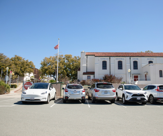 Parking lot with a row of five white and silver of varying models of cars outside of the Veterans Memorial Center Museum.