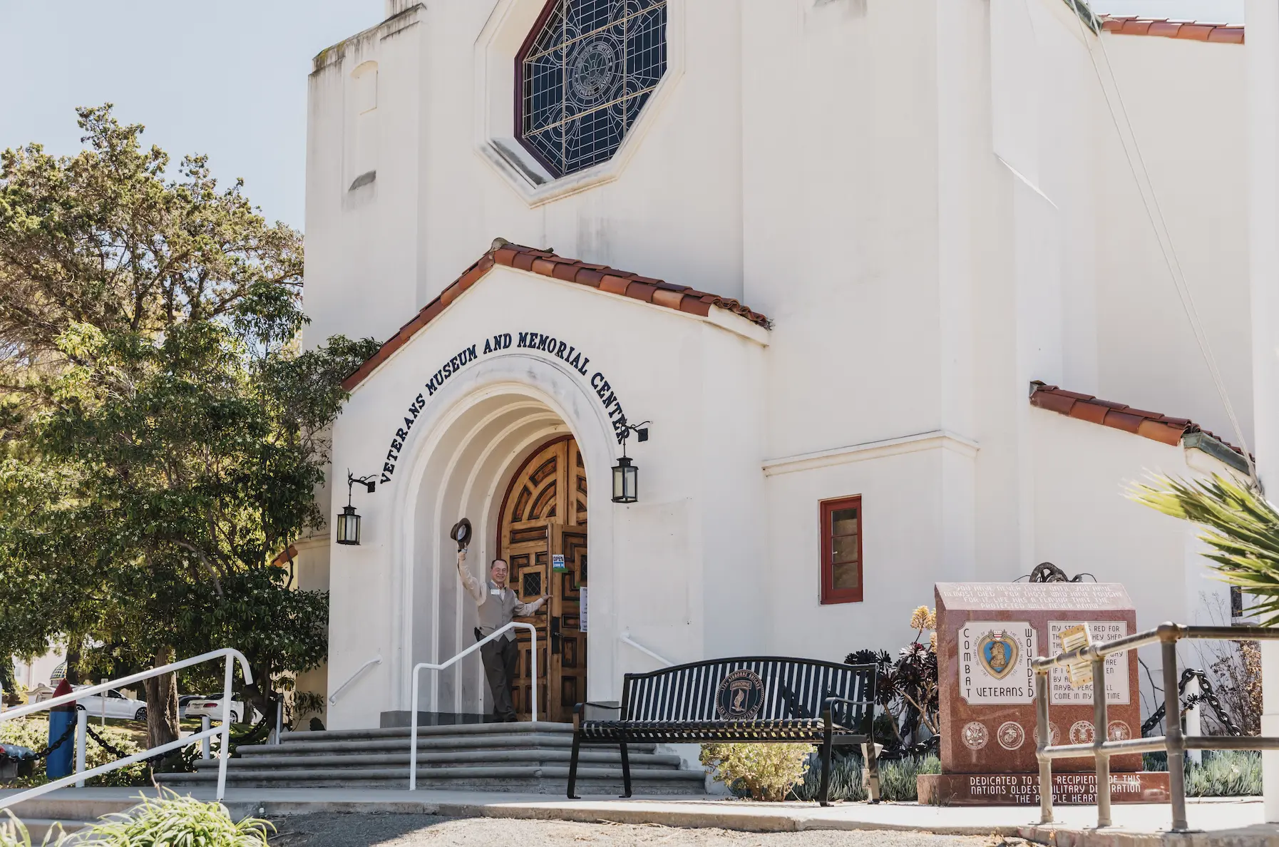 The entrance to the Veterans Museum at Balboa Park. The building is white with a red terracotta roof. The front entry way is arched with wooden doors.