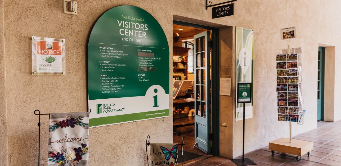 The entry way to the Balboa Park Visitors Center with a green and white arched sign on the left and a display of post cards on the right.