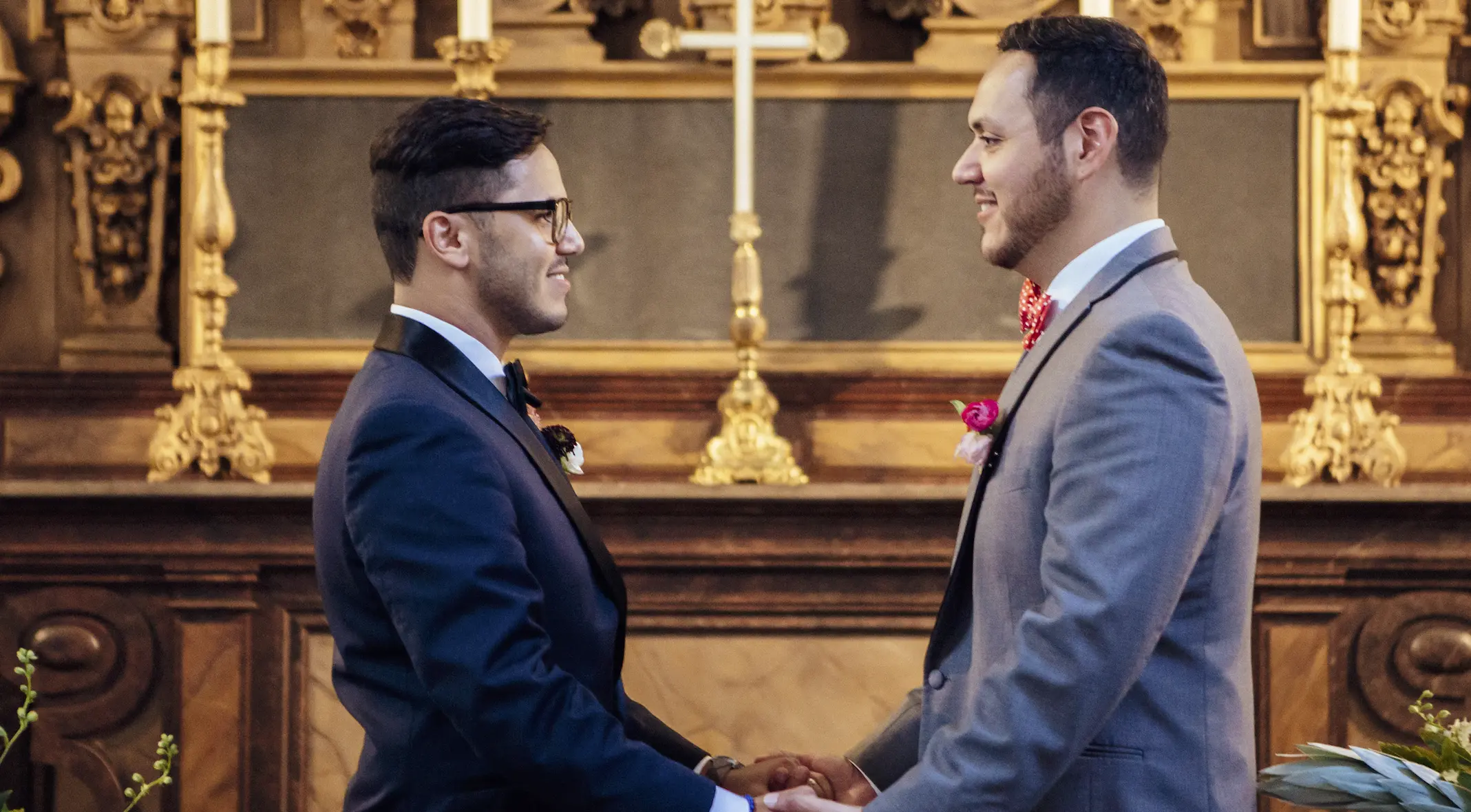 Two adults with brown hair are smiling and facing each other while holding hands. In the background appears to be an ornate gold mantle with candlesticks sitting on top.